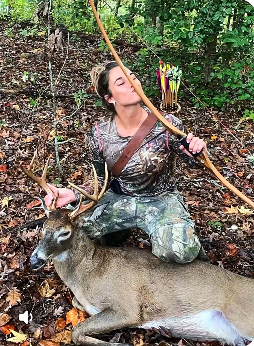 A photo of a woman kneeling behind a white-tailed buck. She's holding up a traditional bow and kissing it, with a quiver of arrows on her back.