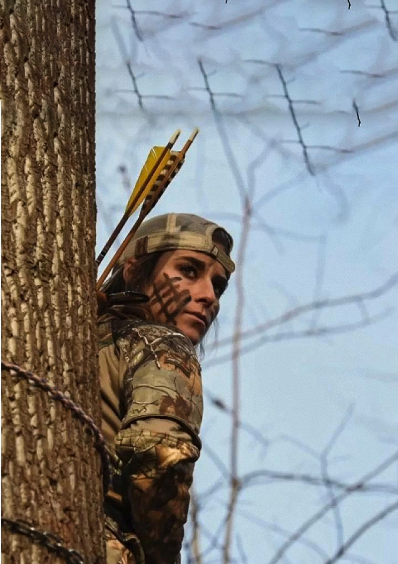 A photo of a woman sitting in a tree stand with traditional-type arrows.