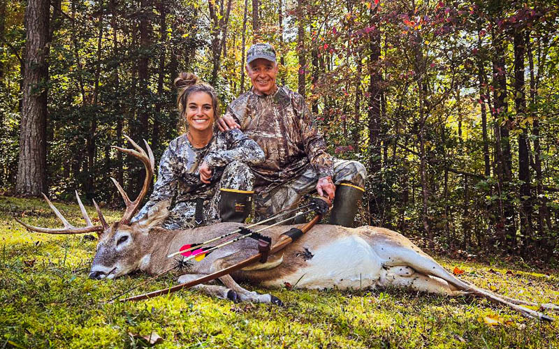 A photo of a woman and man kneeling behind a large white-tailed buck lying on the ground, with traditional archery equipment leaning against the deer.