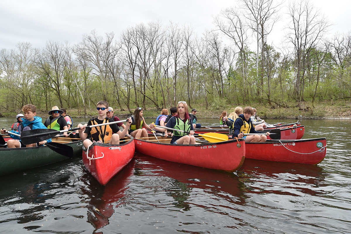 An image of multiple students in red and green canoes and yellow and orange life vests on the water of the James river.