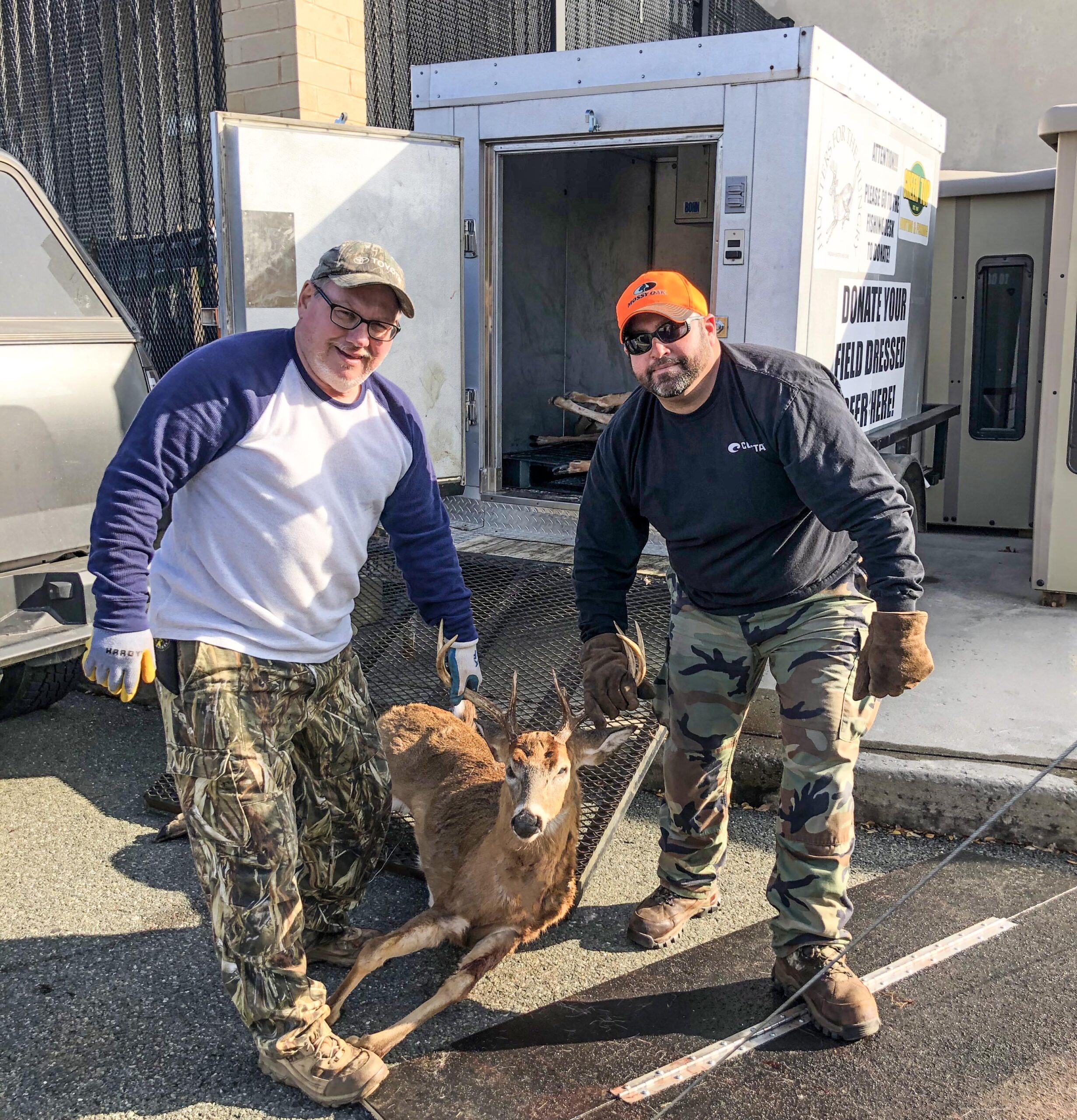 A photo of two men holding up the antlers of a harvested deer in front of a refrigerated trailer.