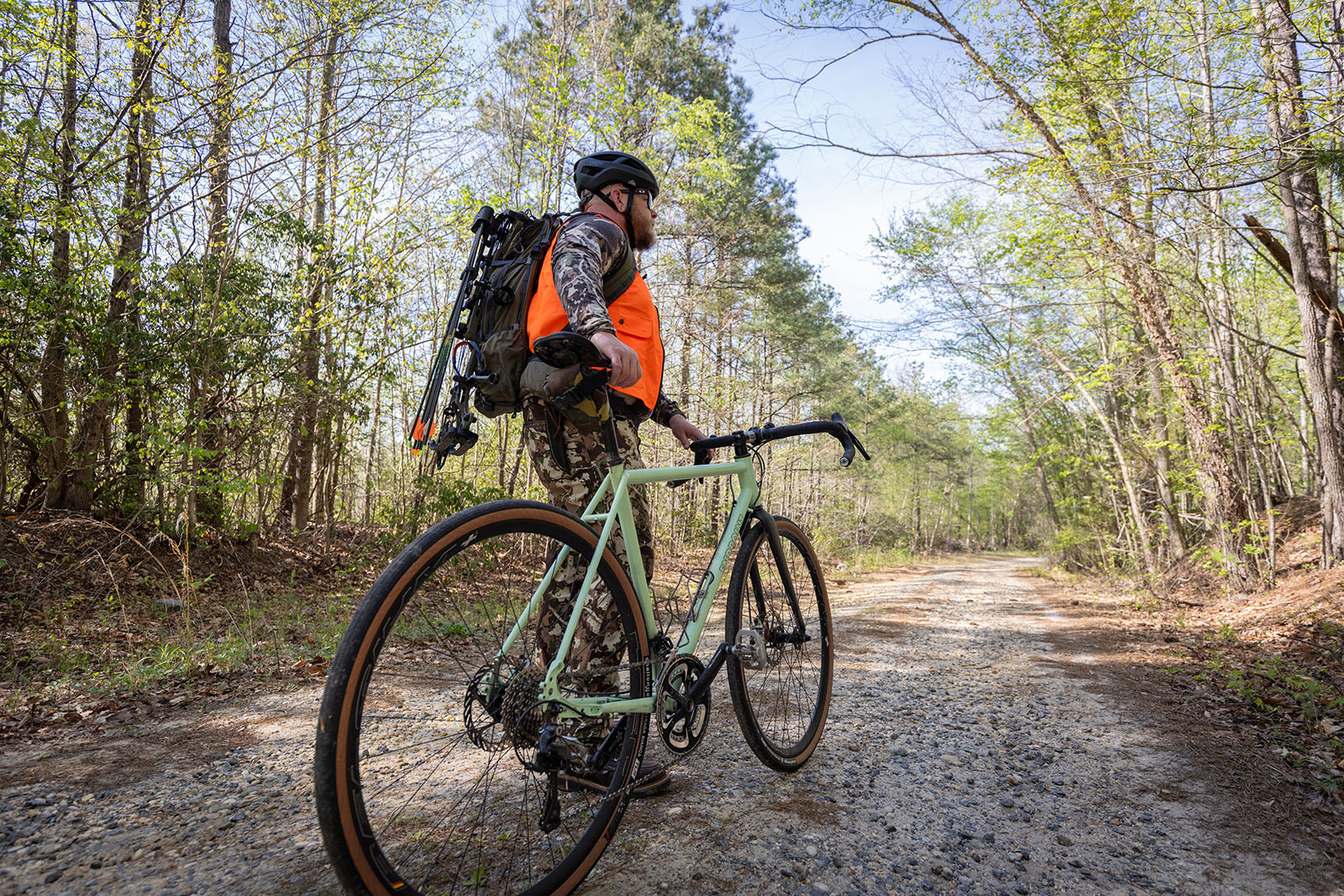 A photo of a man in camouflage and blaze orange, wearing a bike helmet and with a large backpack, stands next to a bike on a gravel road in the woods.
