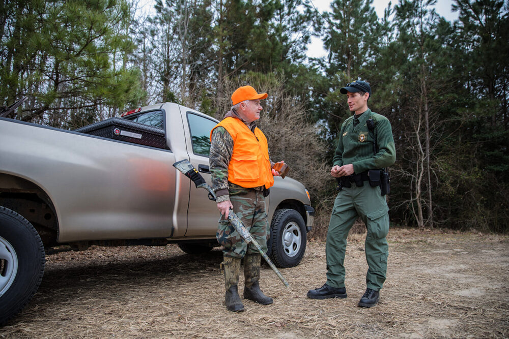 An image of a hunter with a truck and an orange visibility vest talking to a man in green who is a conservation police officer