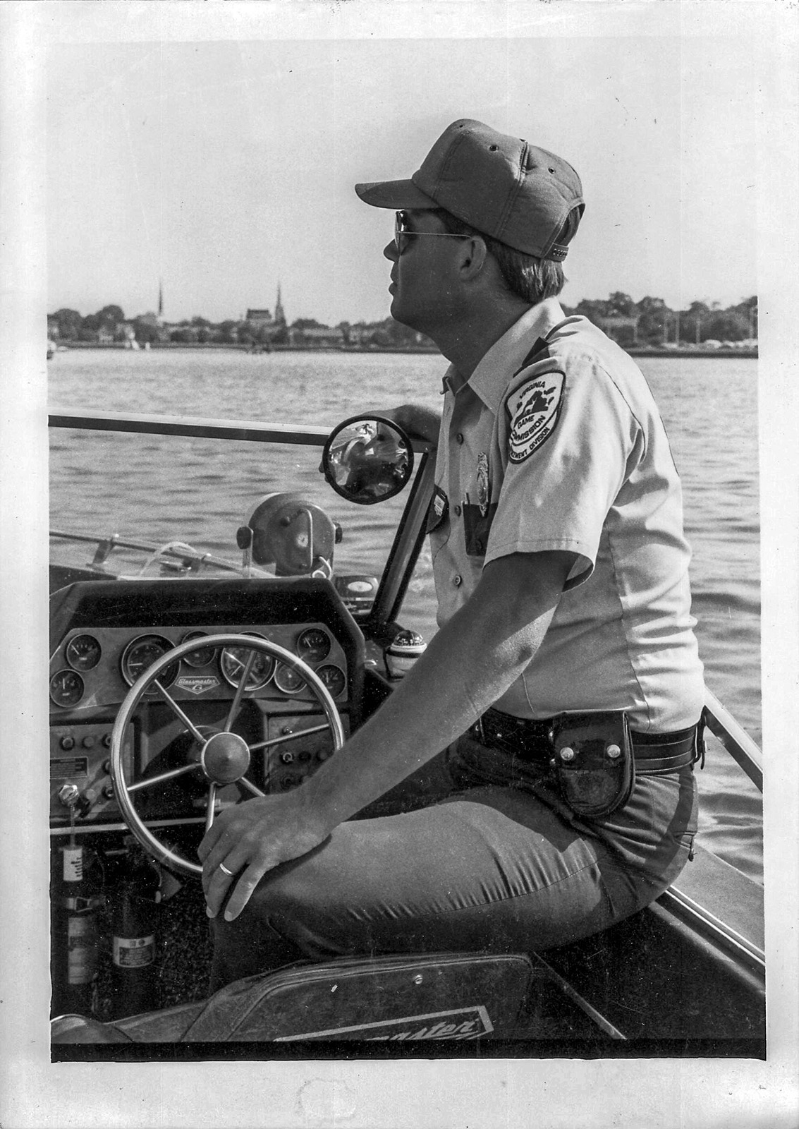 A black-and-white photo of a man in game warden uniform piloting a small boat.