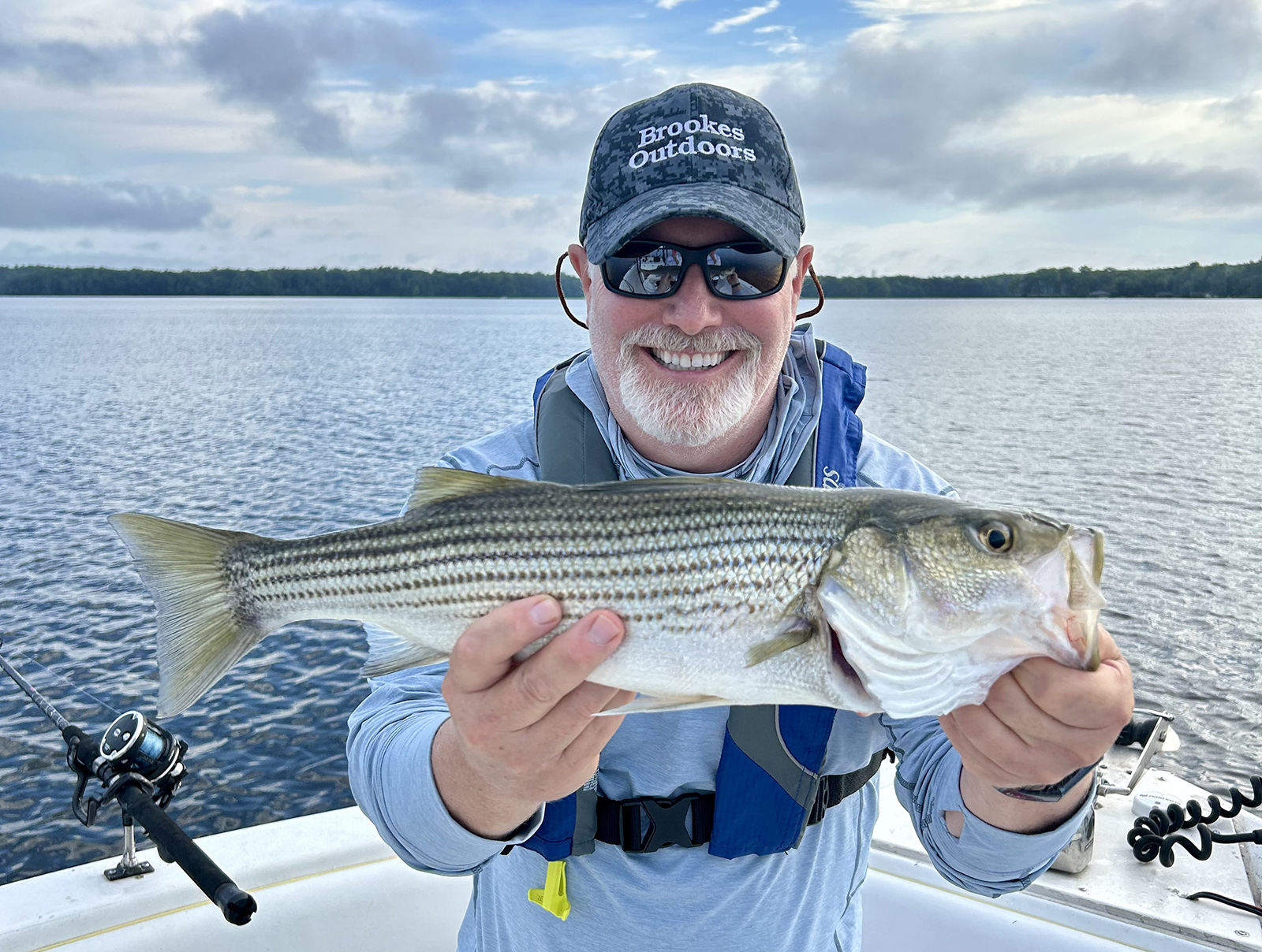 A photo of a smiling man wearing a life jacket and holding a large striped bass on a boat with a lake in the background.