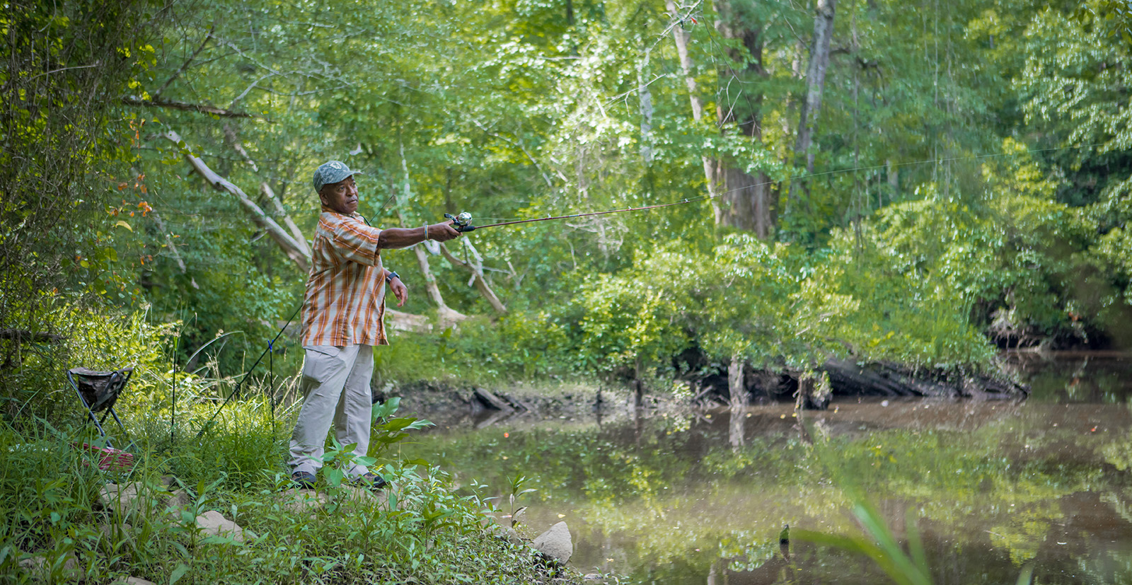 A photo of a man at the edge of a pond, casting a rod and reel into the water.