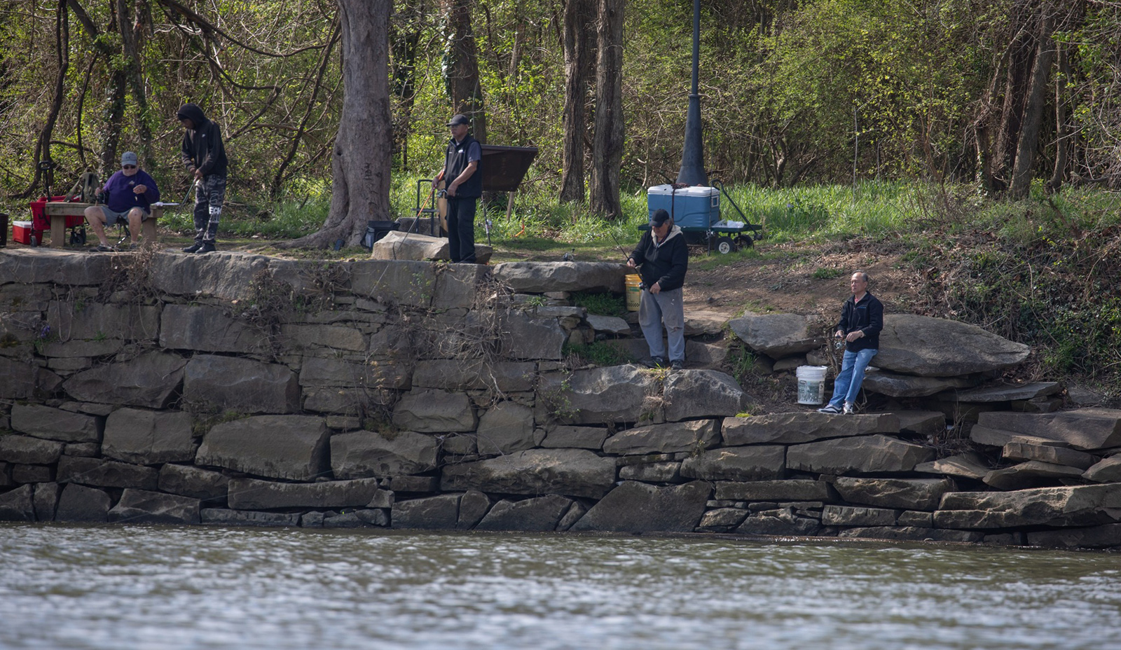 A photo of people standing and sitting on a riverbank and fishing.