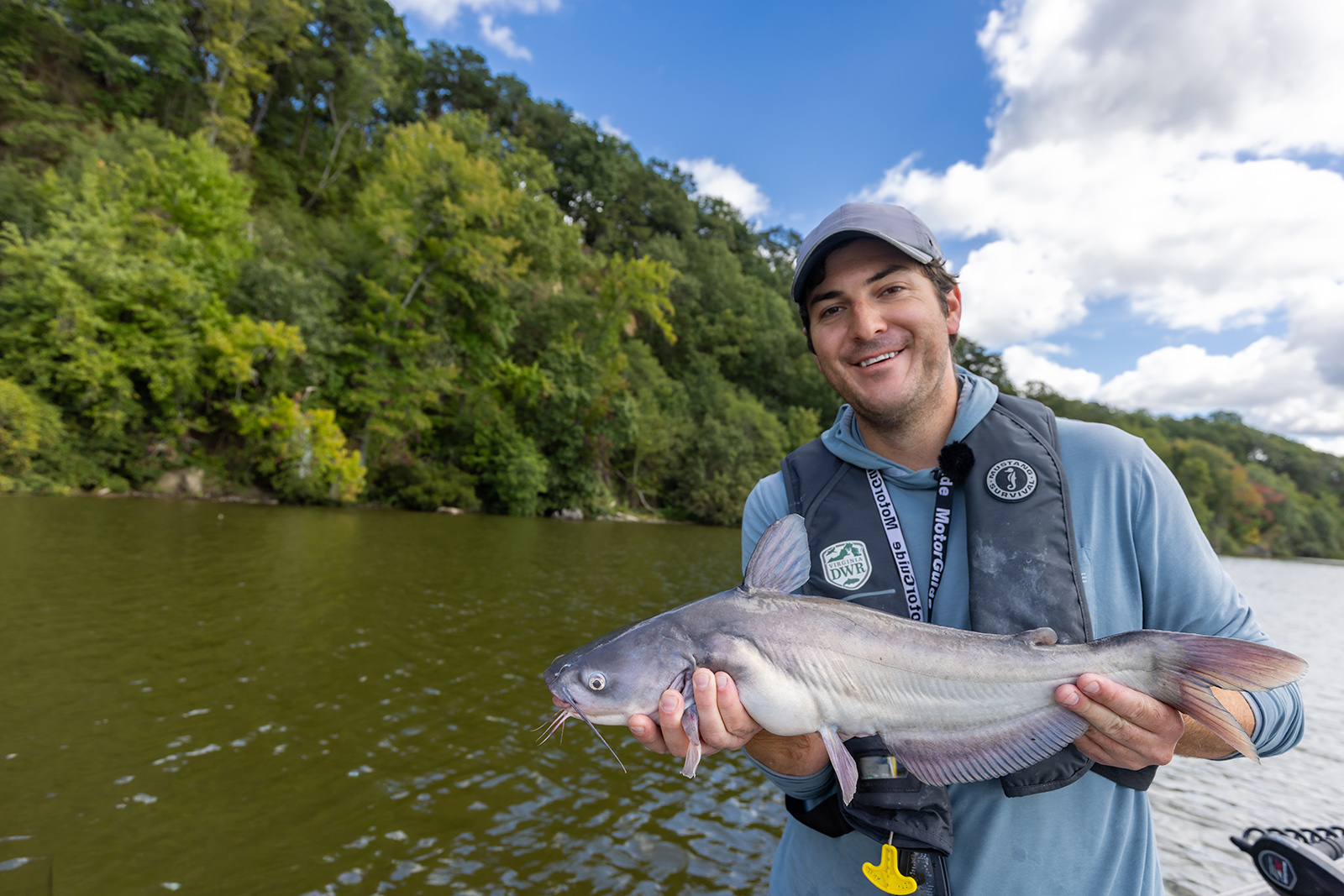 A photo of a man in a life jacket on a boat smiling and holding a medium-size blue catfish.