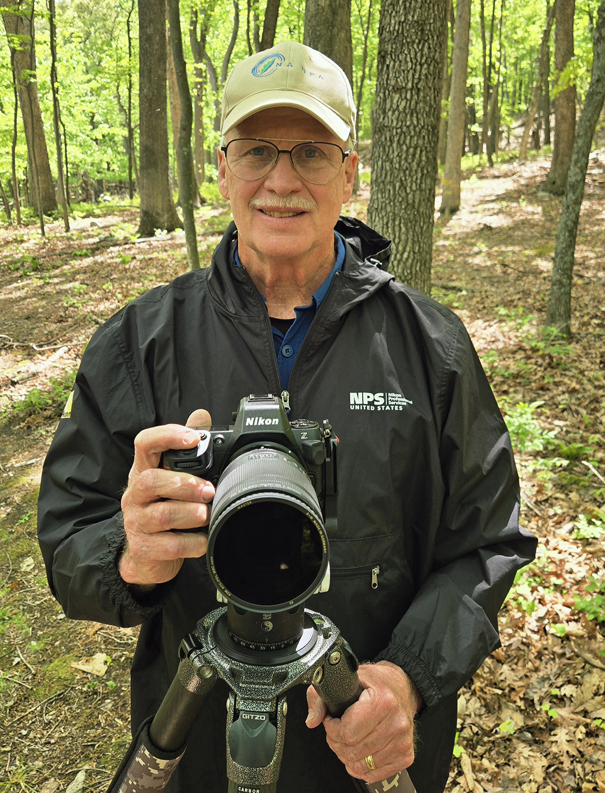A photo of a man holding a camera attached to a tripod while standing in the woods.