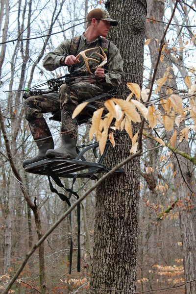 An image of a hunter standing atop a tree platform holding a deer antler; he is clacking them together in attempt to attract bucks