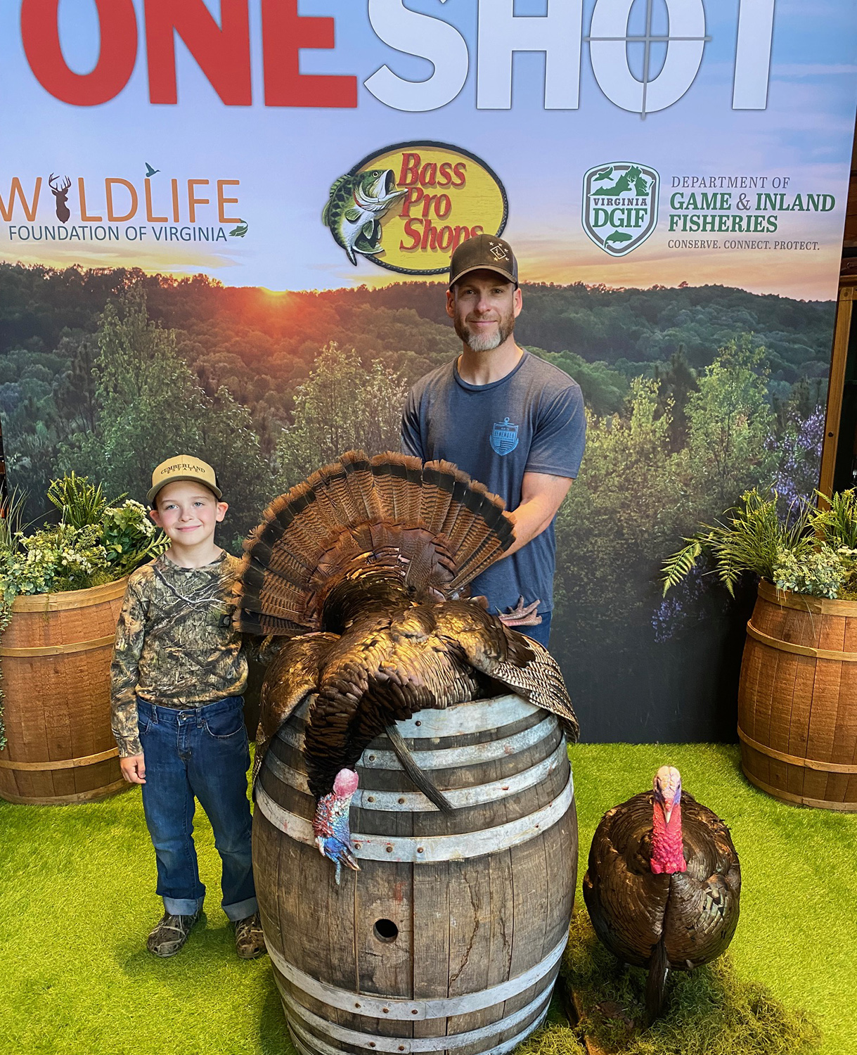 A photo of a young boy and a man posing with a dead wild turkey on top of a barrel and all in front of a backdrop that says One Shot.