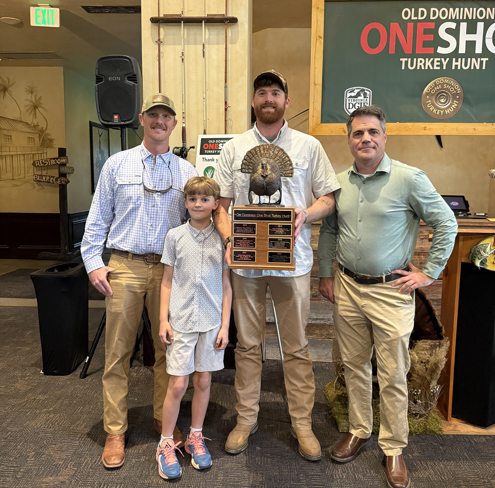 A photo of three men and a boy posing with a large trophy with a turkey on top of it.