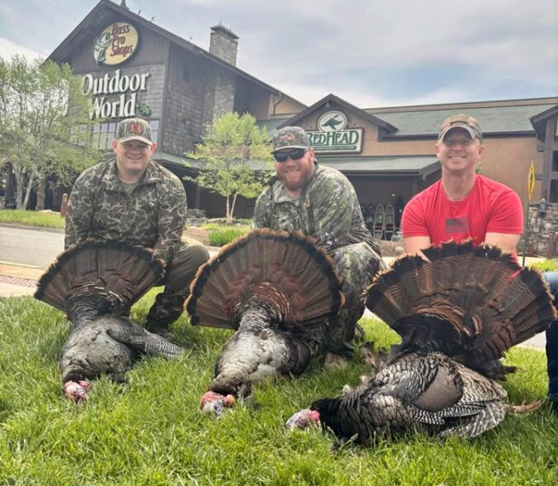 A photo of three men and three dead turkeys on the grass in front of a Bass Pro Shop store.