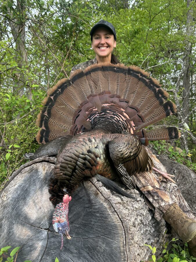 A photo of a female hunter smiling behind a large male turkey laid on a log.