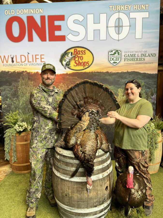 A photo of a man and a woman standing in front of a backdrop with One Shot and sponsor names with a large turkey displayed.