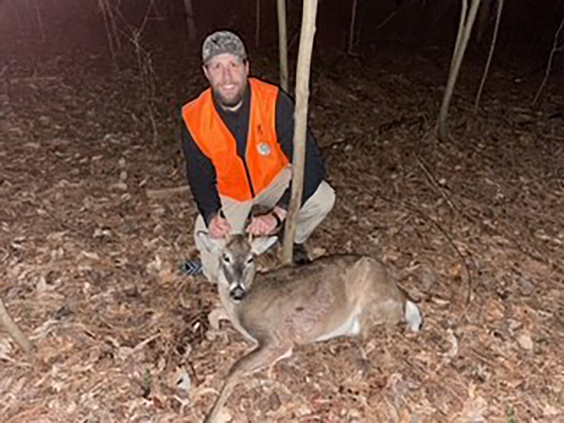 A photo of a man in a blaze orange vest kneeling behind a small whitetailed deer on the ground.