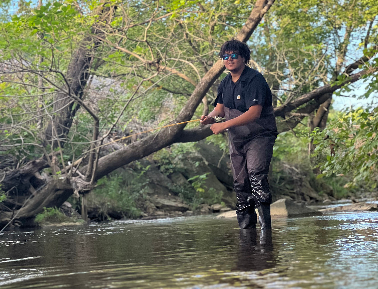 A photo of a man in waders standing ankle-deep in a stream with a fishing pole.