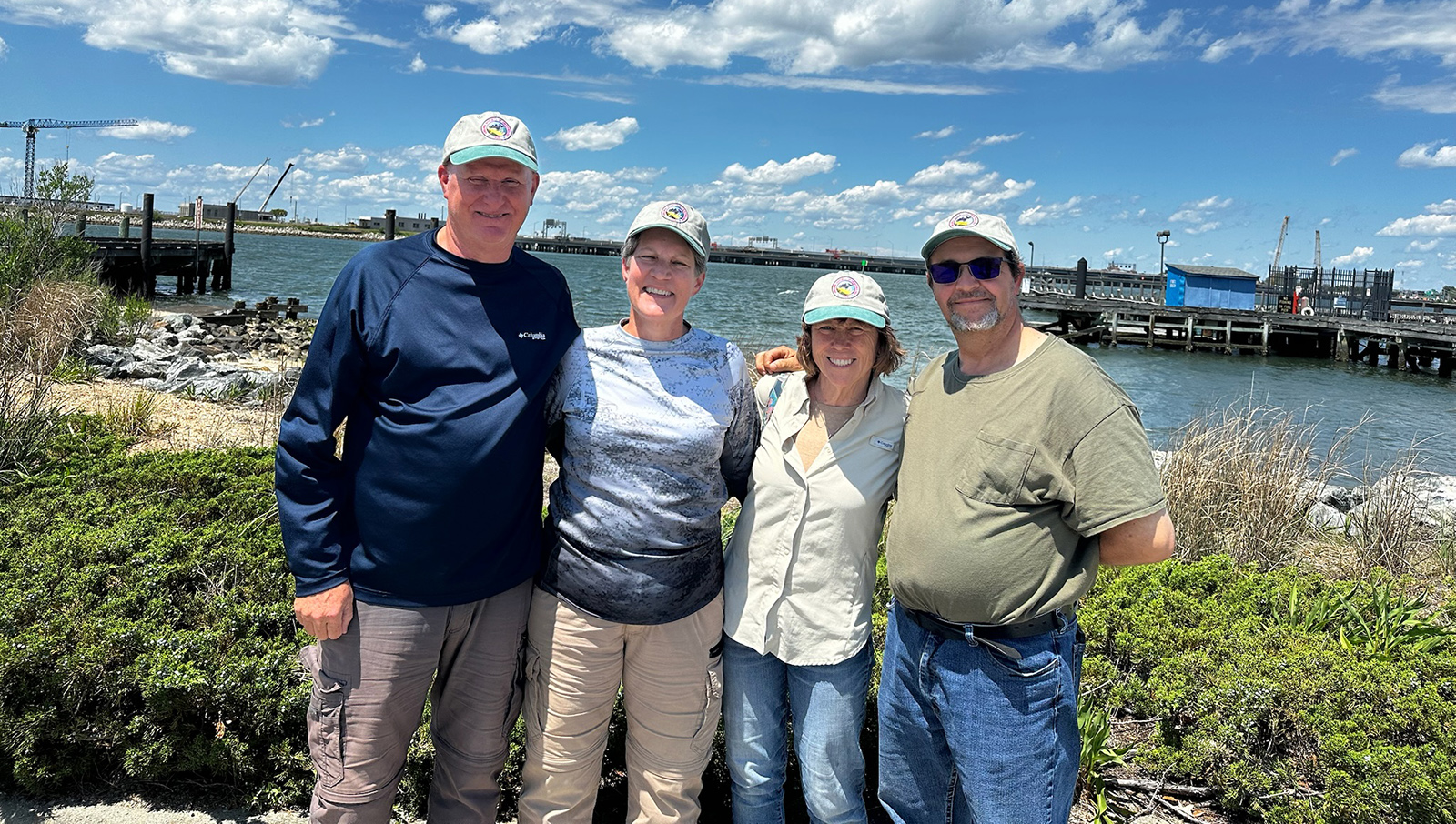 A photo of four smiling people standing on a beach of an island with water in the background.