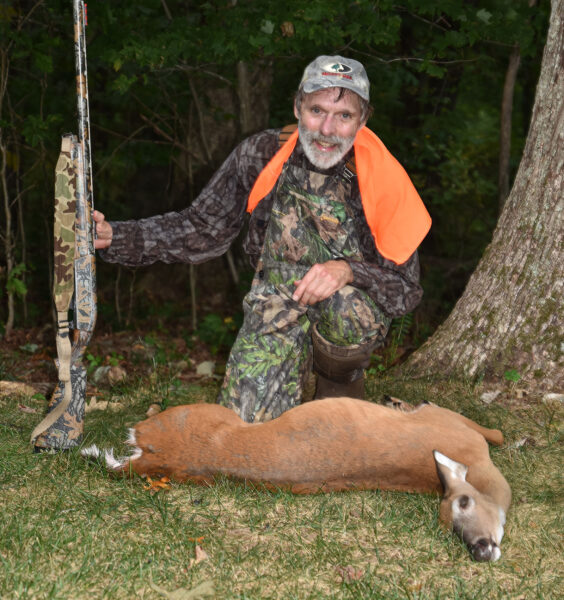 A photo of a man in camouflage and orange holding a gun and kneeling behind a whitetailed doe on the ground.