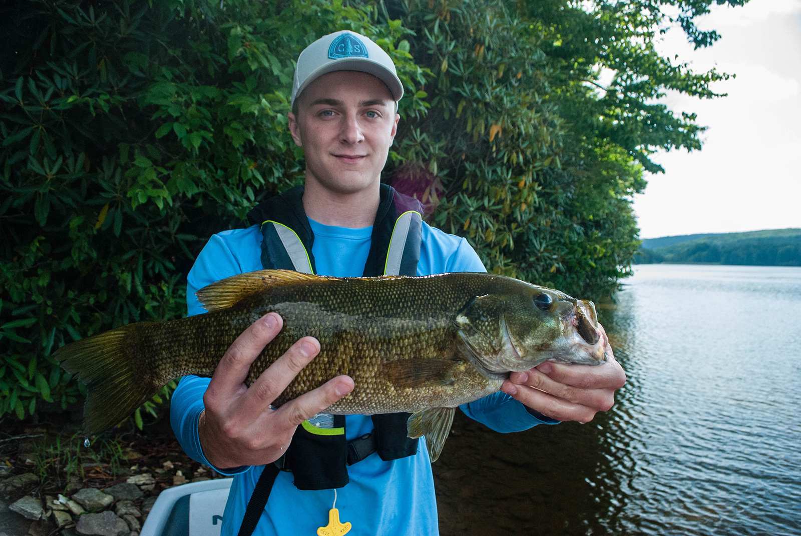 A photo of a young man wearing a life jacket and holding a large smallmouth bass with a lake in the background.