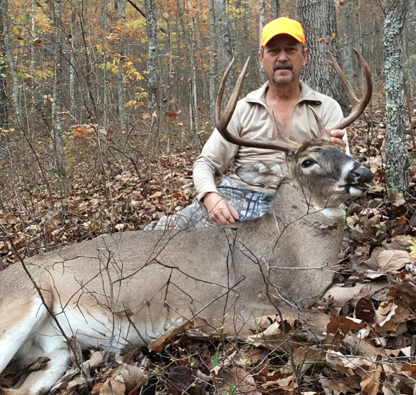 A photo of a man kneeling behind a large whitetail deer buck that's lying on the ground after he shot it.
