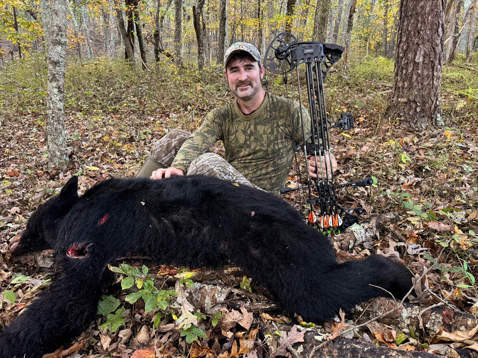 A photo of a man wearing camouflage sitting in the woods next to a dead black bear and holding a crossbow.