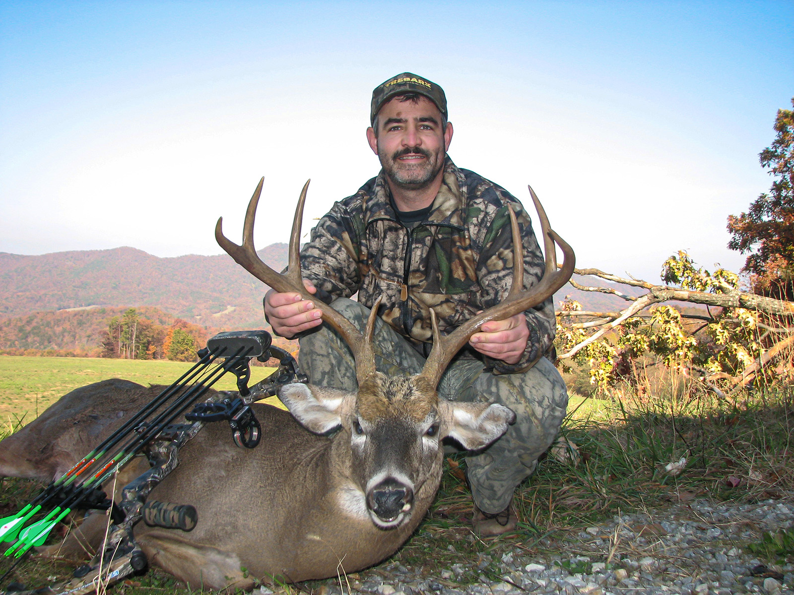 A photo of a man in camouflage kneeling behind a dead, large whitetail deer buck, holding its antlers up.