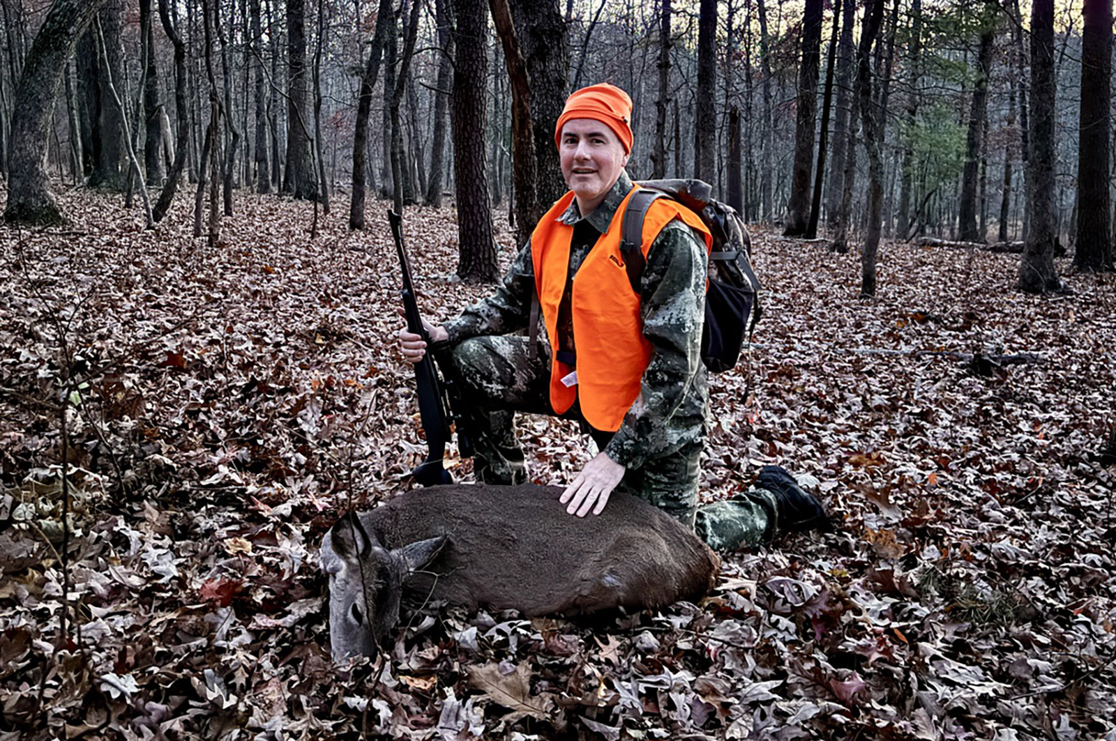 A photo of a man in camouflage and blaze orange kneeling behind a harvested doe deer in the woods.