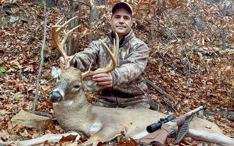 A photo of a man wearing camouflage kneels behind a harvested deer with large antlers.