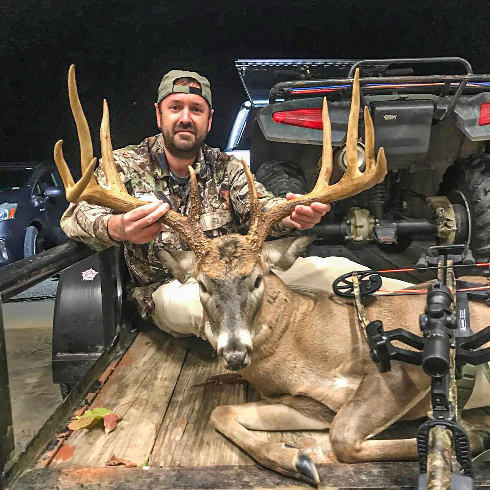 A photo of a man in camouflage in the bed of a truck holding the head of a large buck with 10-point antlers, with a crossbow leaning over the deer's body.