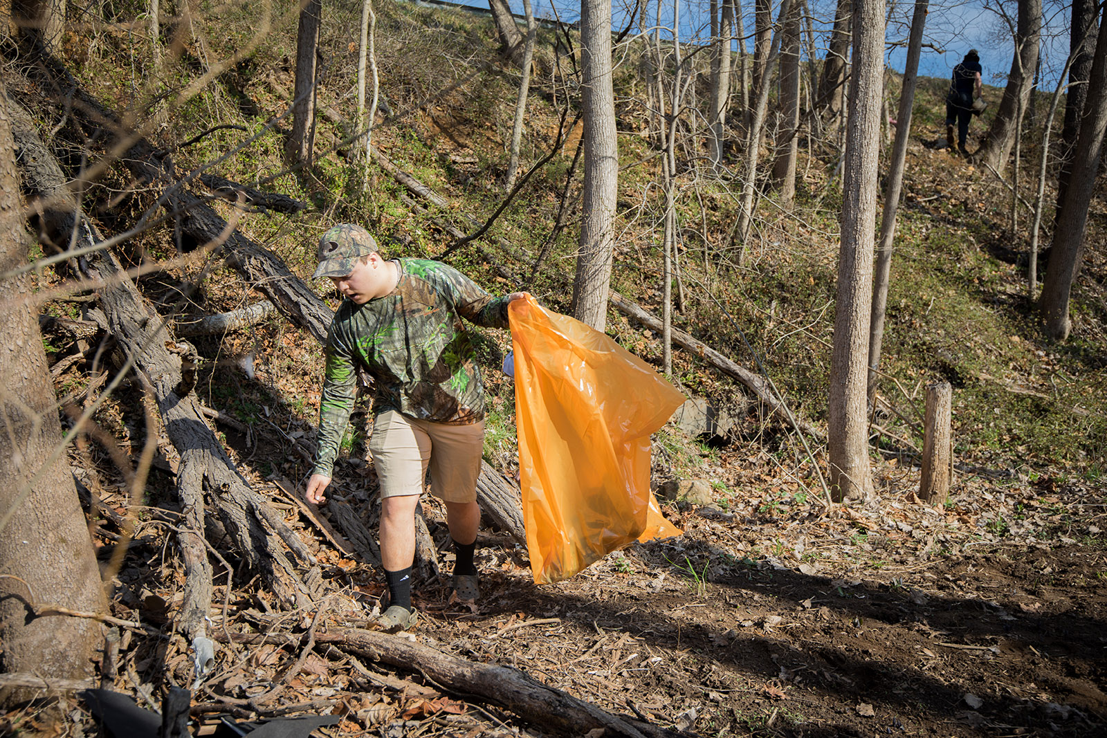 An image of a man in camouflage with a bright orange trash bag picking up garbage in a forest 