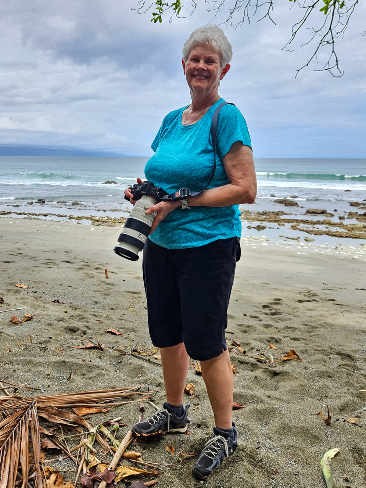 A photo of a woman holding a camera and smiling on a beach.