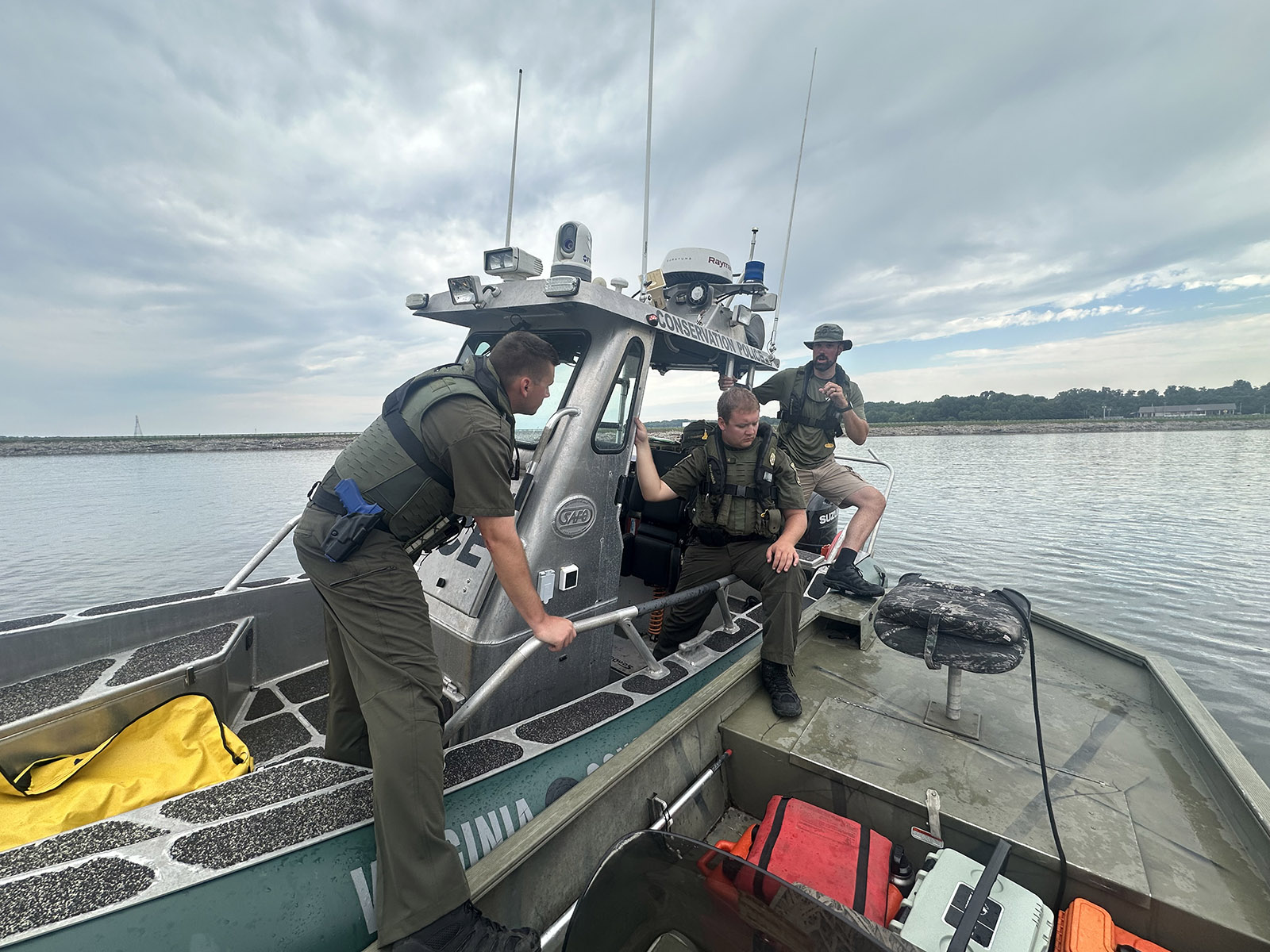 A photo of three men in uniform and life jackets stepping from a Conservation Police boat onto a smaller boat.