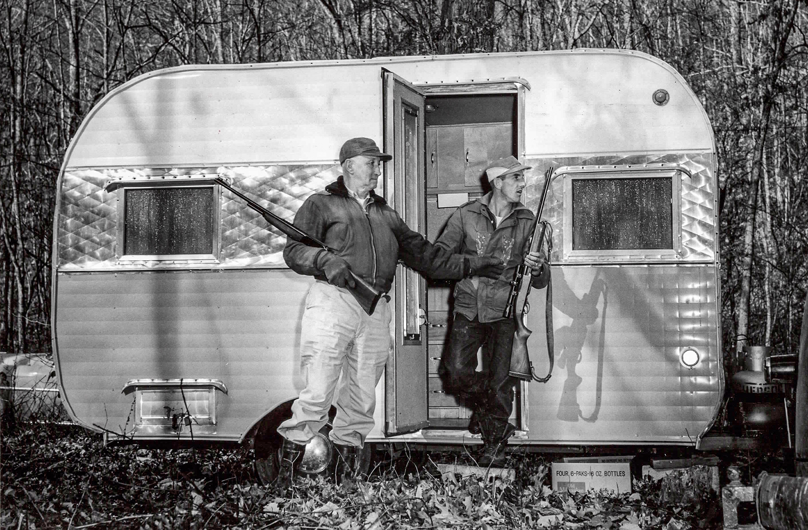 A black and white photo of two men with rifles leaning against a travel trailer in the woods.