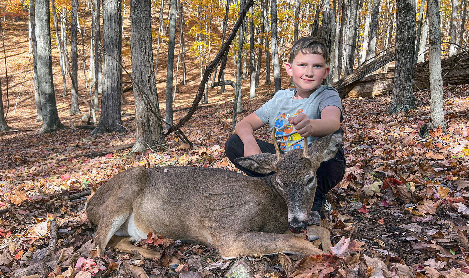 A photo of a young boy sitting behind a small buck white-tailed deer in the woods. 