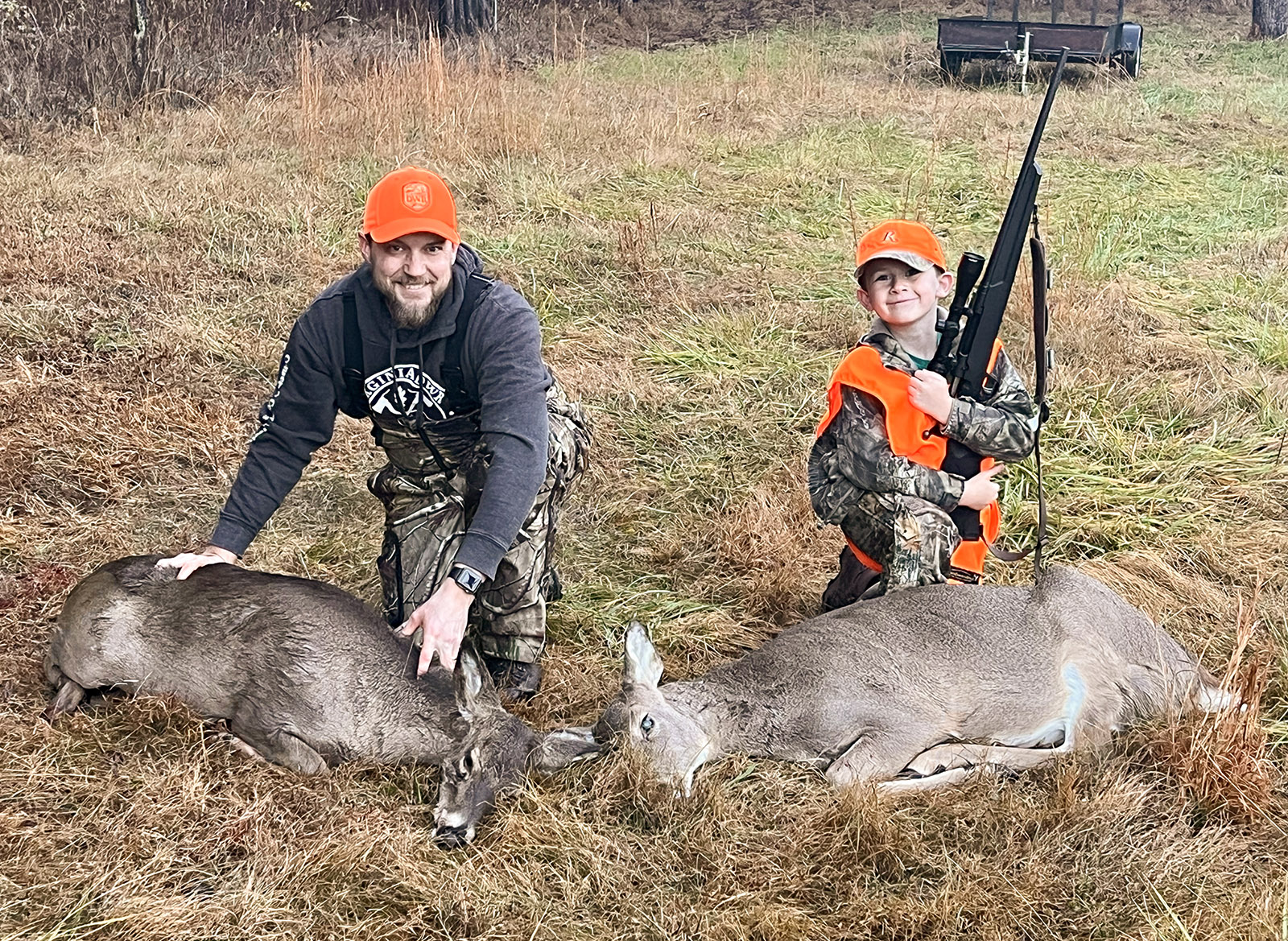 A photo of a man and a boy kneeling behind two harvested doe deer in a field.