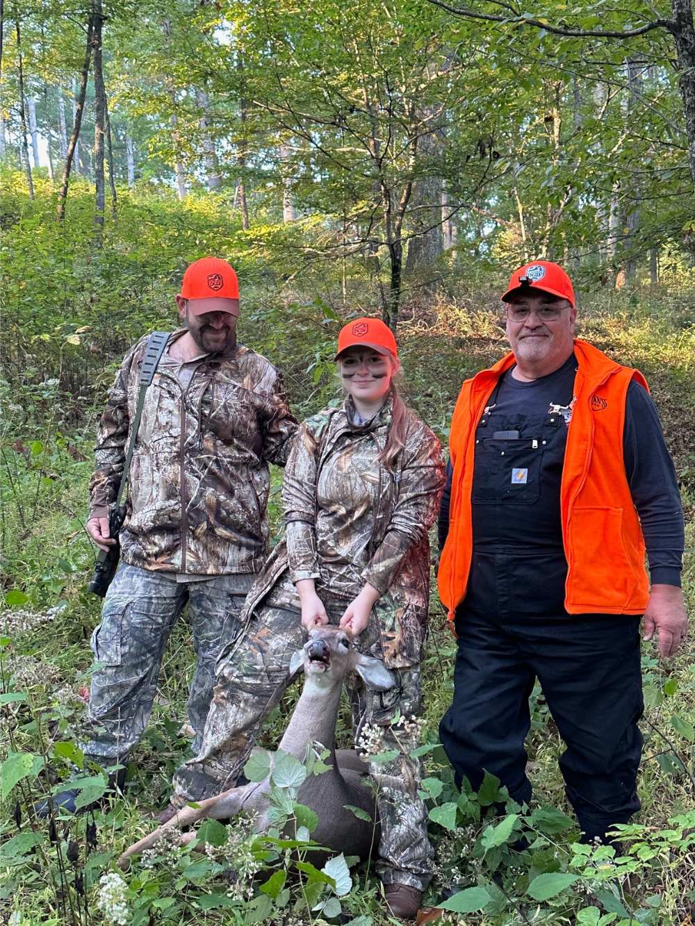 A photo of a woman holding the head of a harvested deer along with two men beside her. They're in the woods and all dressed in camuflage and blaze orange.