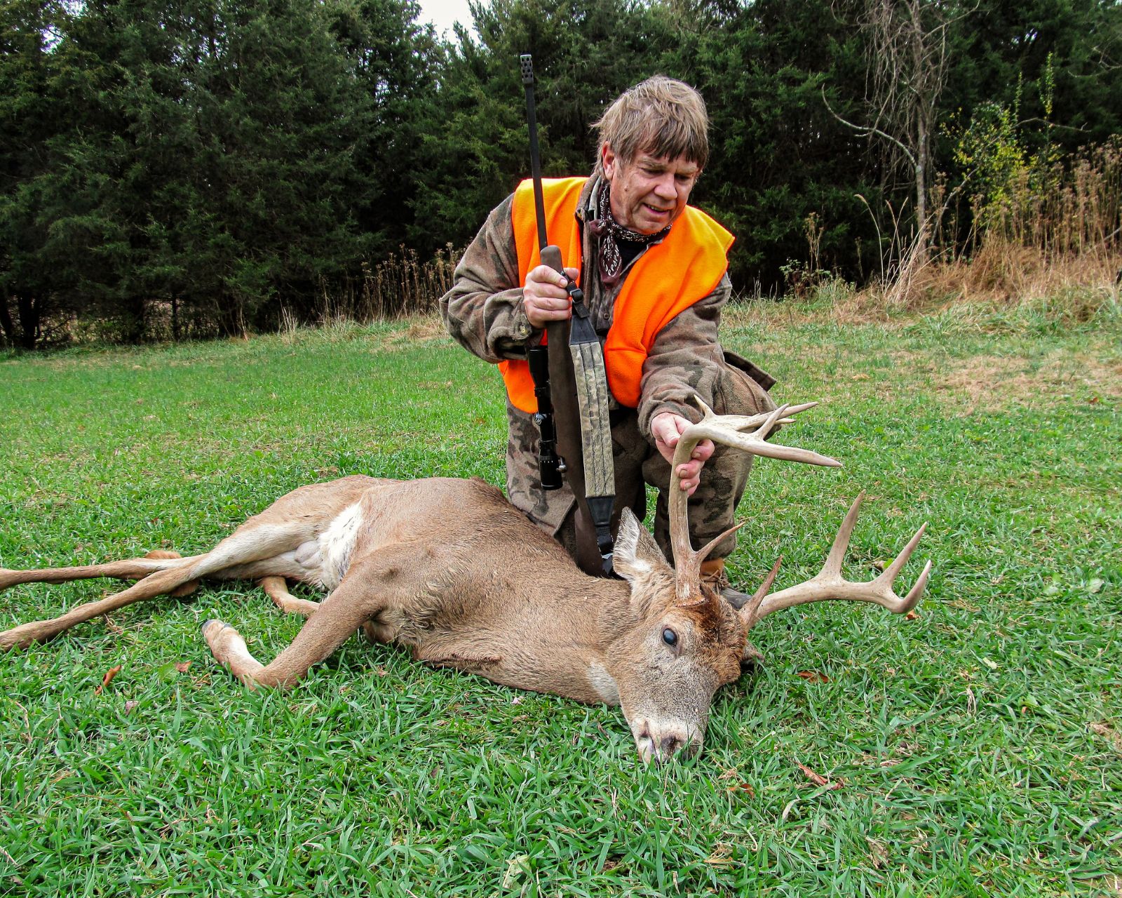 A hunter posing with his gun and a harvested, large, antlered deer. 