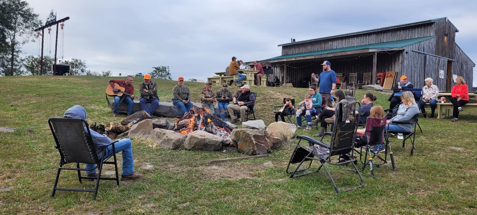 A photo of a campfire with many people seated around it with a barn in the background.