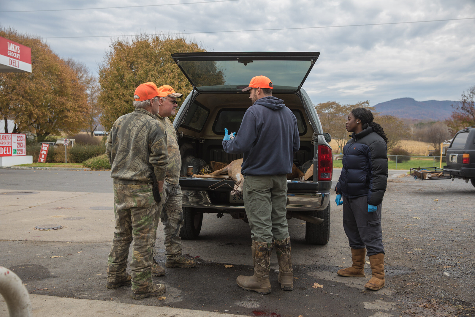 A photo of three men and a woman gathered around a tuck tailgate, where a dead deer is lying, and talking.