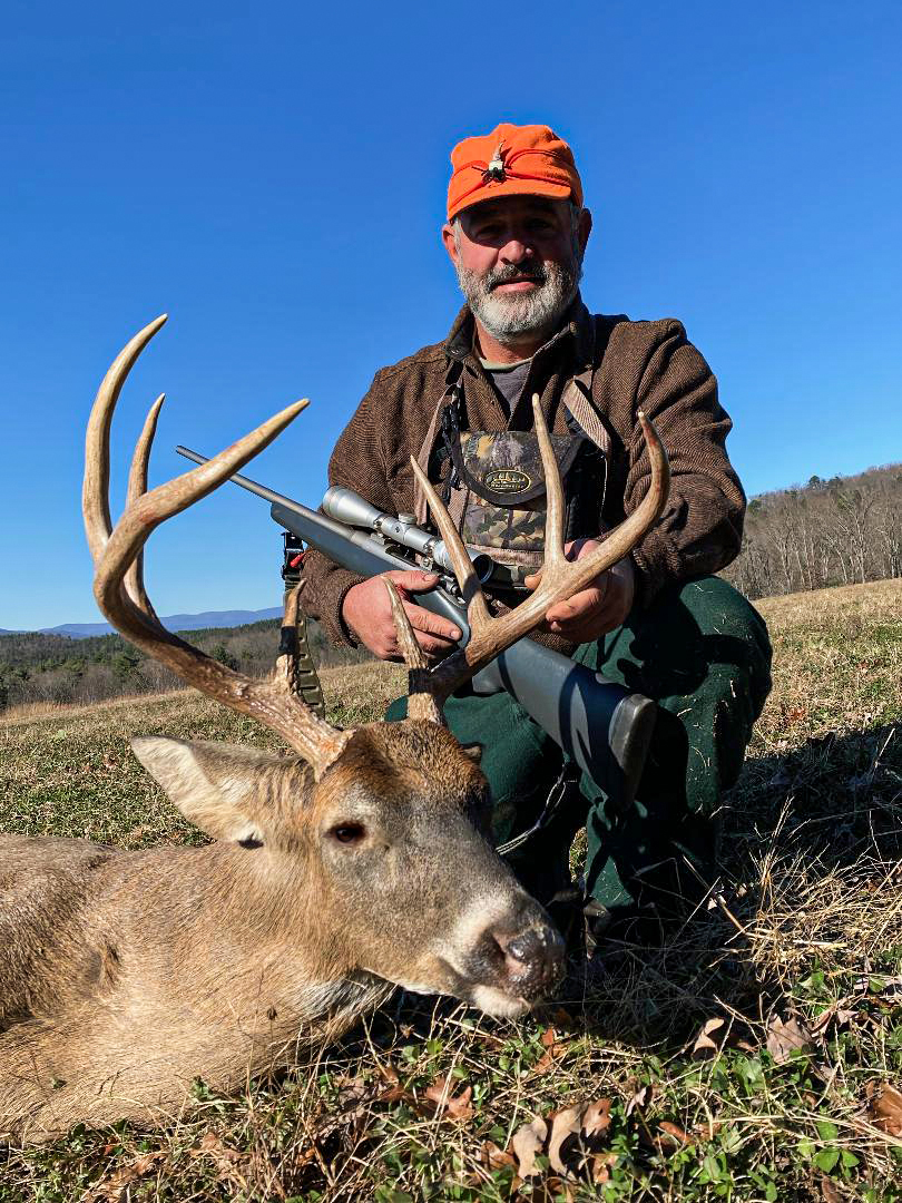 A photo of a large whitetail deer buck laying on the ground with a man kneeling next to it with a rifle, holding the antlers.