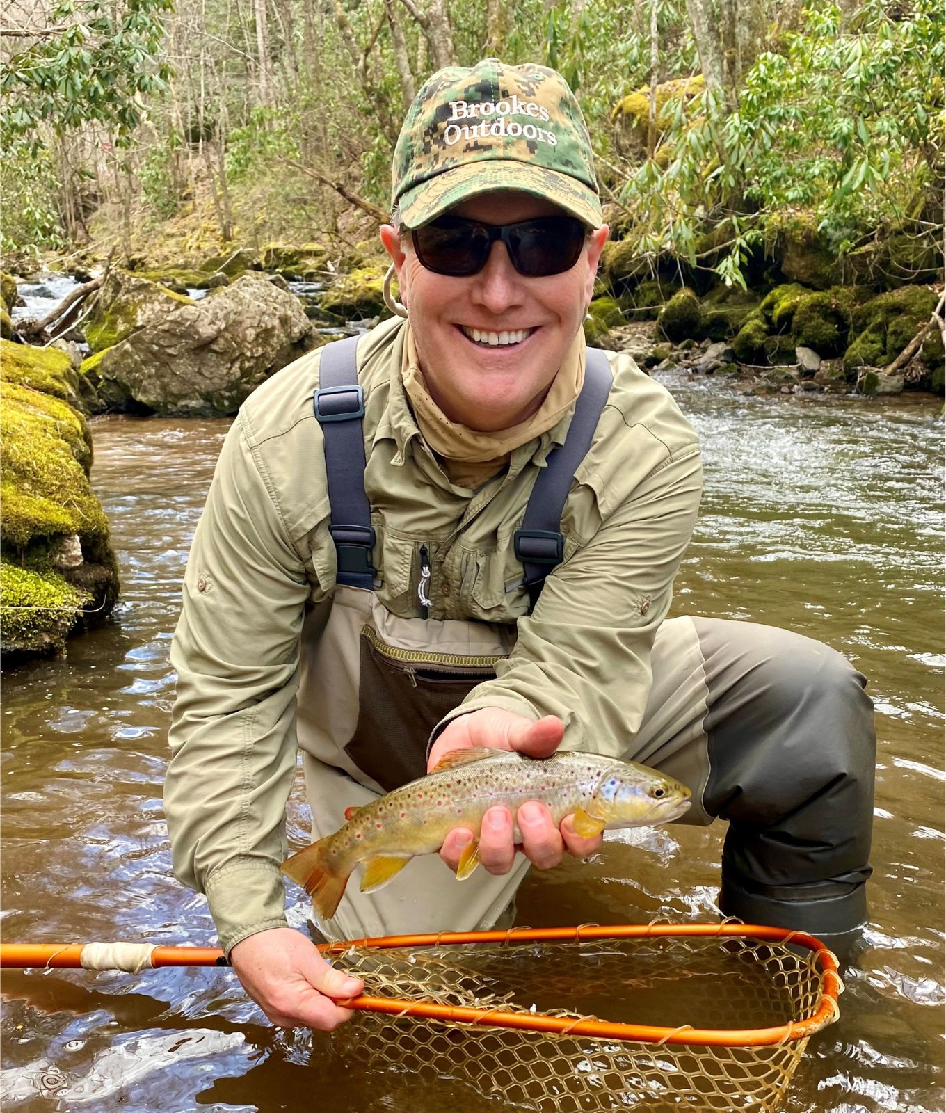 A photo of a smiling man squatting in a beautiful stream, holding a fishing net and a colorful brown trout. 