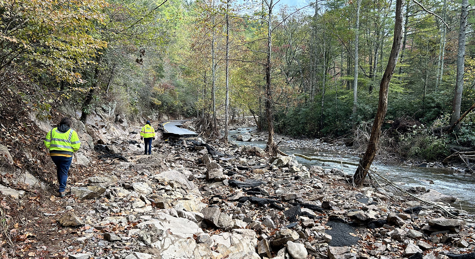 A photo of two people wearing reflective jackets walking a destroyed roadway next to a stream.