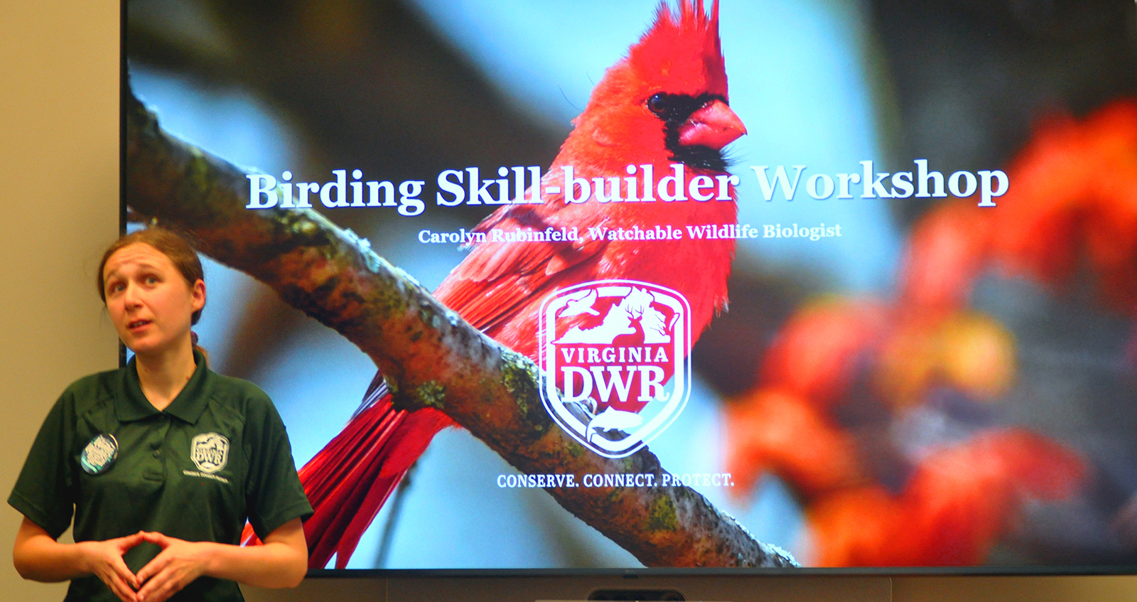 A photo of a woman wearing a DWR-logo shirt in front of a large screen showing a cardinal and the title "Birding Skill-builder Workshop."