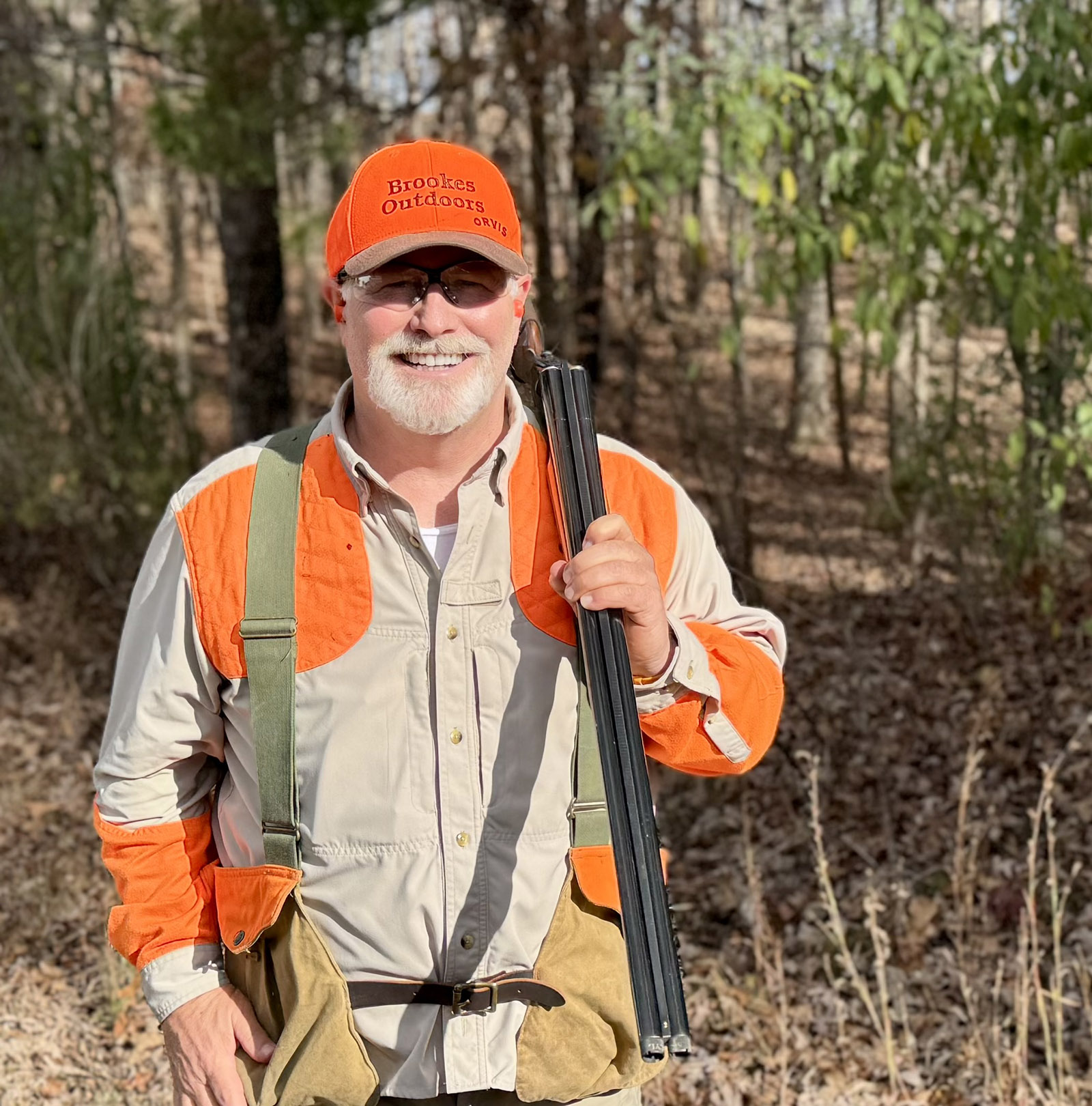 A photo of a man with a shotgun over his shoulder, standing in the woods wearing a blaze-orange hat and a shirt with blaze-orange patches on the shoulders and arms.