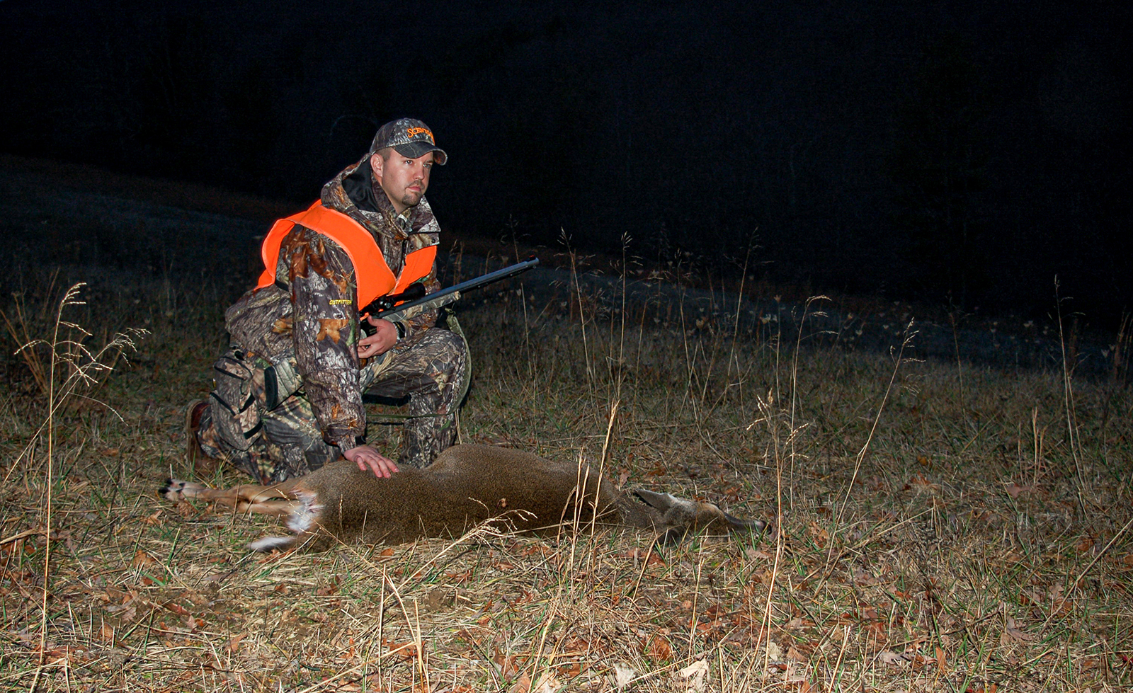 A hunter with gun and dressed in camouflage kneels next to a deer lying on the ground in the dark.