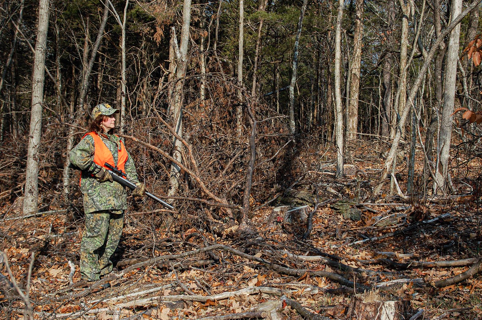 A photo of a hunter holding a gun, dressed in camouflage and blaze orange, standing among cut trees in a forest.