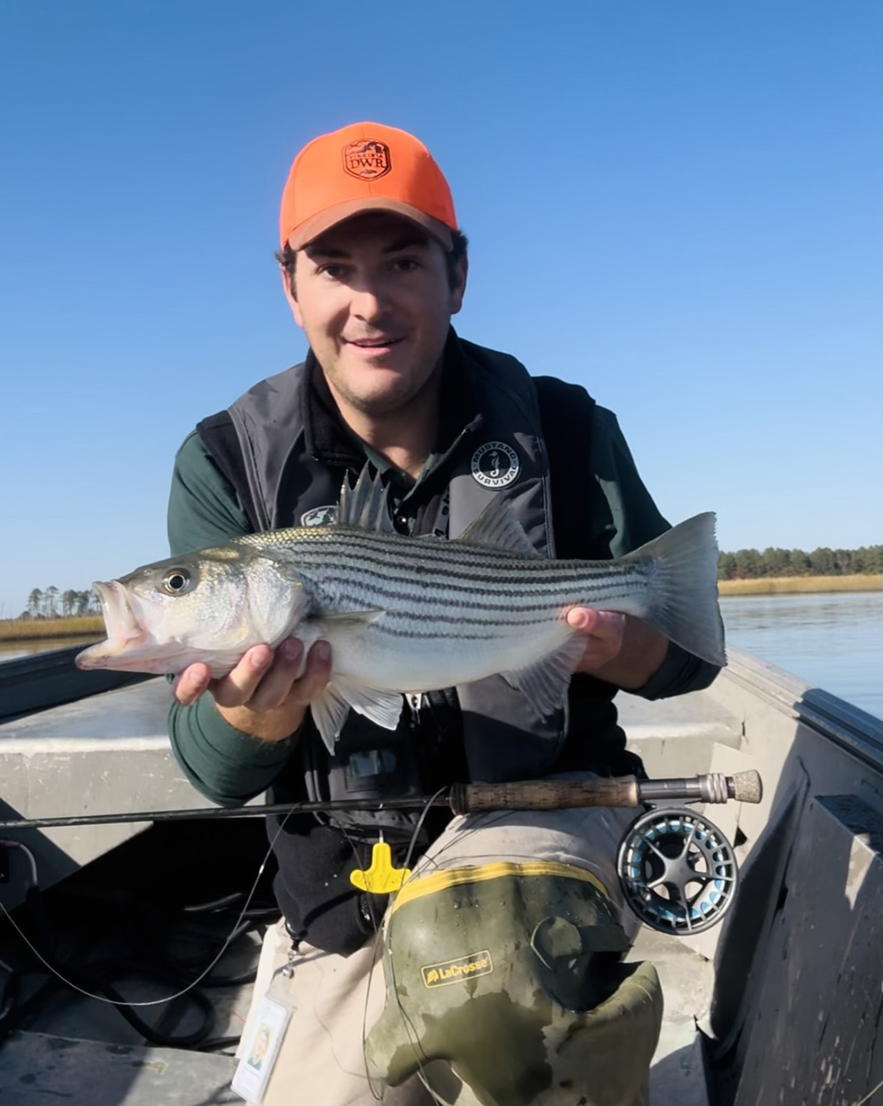 A photo of a man kneeling at the front of a small boat, holding up a striped bass he caught.