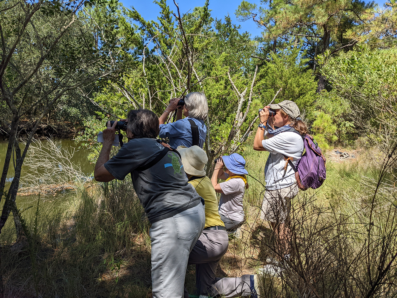 A photo of a group of people on the banks of a river, looking through binoculars across the water.