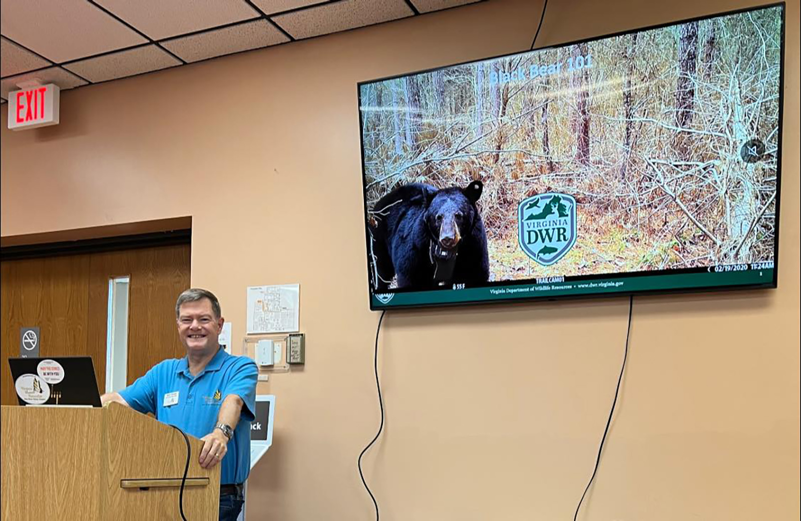 A photo of a man behind a lectern giving a presentation, with a photo of a black bear on the screen behind him.